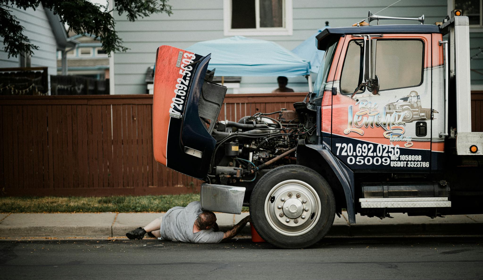 A mechanic works under a truck on a neighborhood street, highlighting vehicle maintenance.